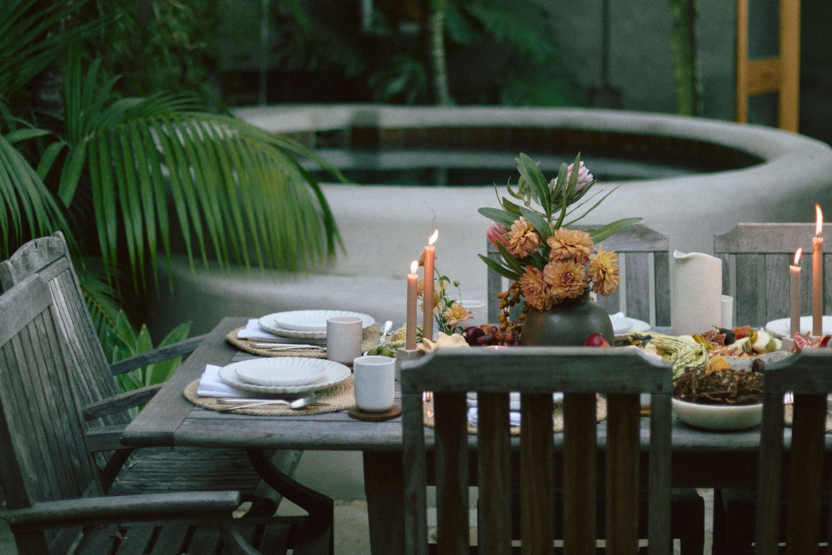 A rustic wooden dining table set with plates, candles, and floral centerpieces in a lush tropical garden courtyard with a hot spring or spa pool in the background.